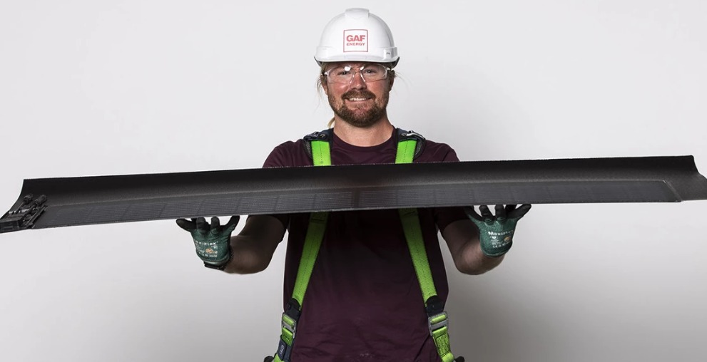 A photo of a person in a hardhat and roofer's harness holding a GAF Timberline solar shingle in front of a white background